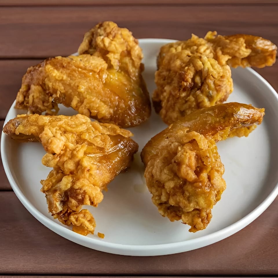 Four crispy, golden-brown fried chicken wings on a white plate, against a wooden table.